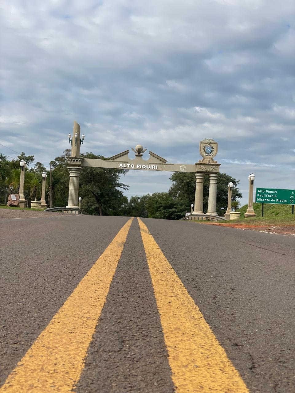 Você está observando o portal de entrada do município de Alto Piquiri, no estado do Paraná. A fotografia foi registrada a partir do centro da rodovia, destacando as faixas amarelas da pista que conduzem o olhar diretamente até o monumento. O portal possui colunas ornamentadas, iluminação decorativa e o nome da cidade em destaque na parte central da estrutura. No lado direito encontra-se o brasão oficial do município, elemento que representa a identidade histórica e administrativa de Alto Piquiri. Ao redor do portal, observam-se áreas verdes, árvores e uma placa de sinalização indicando as distâncias para Alto Piquiri, Paulistânia e Mirante do Piquiri. Além de sua função de recepção aos visitantes, o portal constitui um importante marco simbólico de identificação territorial, representando a história, a cultura e o sentimento de pertencimento da população local. Este registro integra o Guia Virtual do Projeto EPT do Colégio Estadual Alto Piquiri, produzido em 2026.