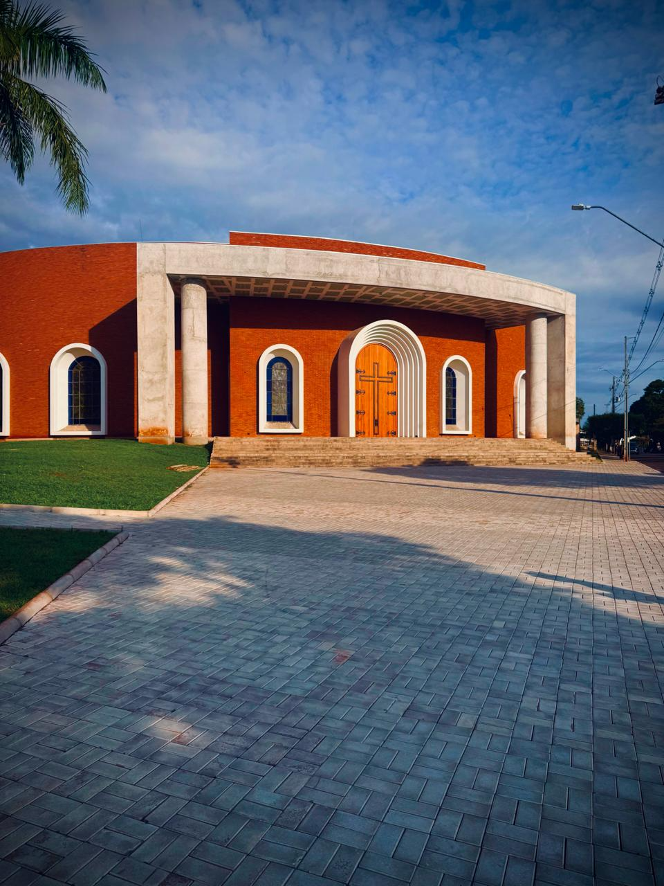 A fotografia apresenta a fachada principal do Santuário São José, um dos mais importantes patrimônios religiosos, culturais e arquitetônicos do município de Alto Piquiri, localizado na região noroeste do Paraná. A edificação destaca-se por sua arquitetura contemporânea, caracterizada pela combinação de linhas curvas, elementos monumentais e harmoniosa integração entre concreto aparente, revestimentos em tijolo cerâmico e amplas áreas abertas para acolhimento dos visitantes. O pórtico frontal, sustentado por colunas circulares de grande porte, valoriza a entrada principal e confere imponência ao conjunto arquitetônico. Ao centro da fachada encontra-se a porta principal em madeira, emoldurada por arcos sucessivos que criam profundidade visual e reforçam o simbolismo da entrada para um espaço de oração, contemplação e encontro comunitário. As janelas laterais em formato arqueado dialogam com elementos tradicionais da arquitetura religiosa cristã, reinterpretados em linguagem contemporânea. A ampla praça pavimentada em frente ao santuário favorece a circulação de fiéis, visitantes e peregrinos, funcionando como espaço de convivência, celebrações religiosas e eventos comunitários. A área ajardinada complementa a composição paisagística, contribuindo para a valorização estética do patrimônio. Além de sua função religiosa, o Santuário São José representa um importante marco da memória coletiva de Alto Piquiri, simbolizando a fé, a identidade cultural e a trajetória histórica da comunidade local. Fonte: Registro fotográfico da equipe do Projeto EPT, 2026.