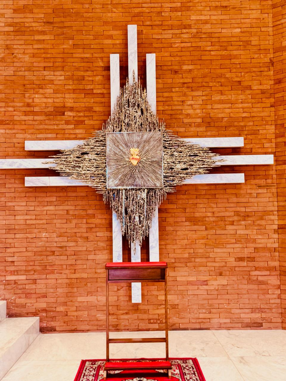 A fotografia apresenta um dos elementos artísticos e devocionais do interior do Santuário São José, em Alto Piquiri, Paraná. No centro da composição encontra-se a representação do Sagrado Coração de Jesus, símbolo tradicional da espiritualidade cristã que expressa o amor, a misericórdia e a compaixão de Cristo pela humanidade. A obra está instalada sobre uma estrutura monumental composta por linhas verticais e horizontais revestidas em mármore claro, formando uma cruz estilizada. Ao centro destaca-se um painel metálico ornamentado, do qual irradiam múltiplos elementos que simbolizam luz, presença divina e expansão da fé cristã. O fundo em tijolos aparentes cria contraste visual com os materiais utilizados na composição artística, valorizando os detalhes do conjunto e reforçando a identidade arquitetônica contemporânea do santuário. Na parte inferior da imagem observa-se um genuflexório utilizado pelos fiéis para momentos de oração, reflexão e devoção. O espaço constitui um importante ponto de contemplação religiosa dentro do templo, favorecendo a experiência espiritual dos visitantes. A obra integra o patrimônio artístico e religioso do Santuário São José, contribuindo para a preservação da fé, da cultura e da memória religiosa da comunidade de Alto Piquiri. Fonte: Registro fotográfico da equipe do Projeto EPT, 2026.