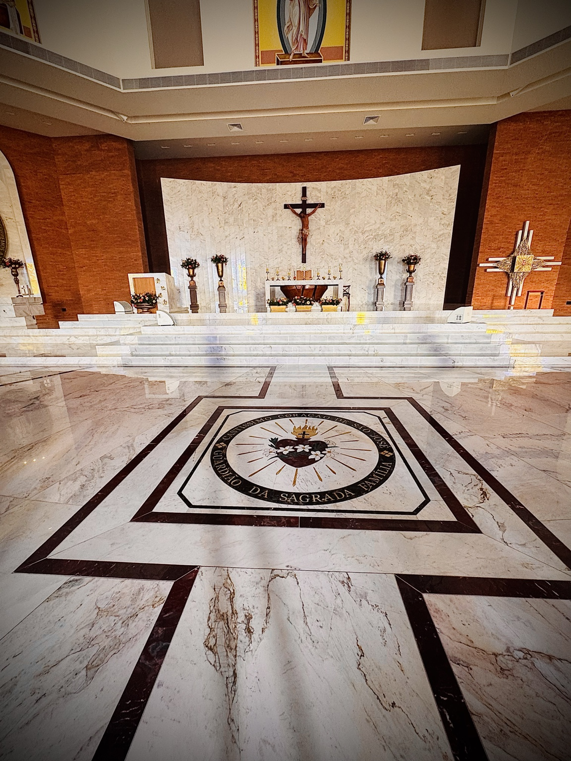 A fotografia apresenta uma vista panorâmica do altar principal do Santuário São José, um dos espaços mais importantes para a celebração da fé e da vida comunitária no município de Alto Piquiri. O registro destaca a amplitude e a harmonia arquitetônica do interior do templo, evidenciadas pelo revestimento em mármore claro, pelos elementos litúrgicos cuidadosamente organizados e pela composição visual que conduz o olhar até o altar central. Ao fundo, observa-se o crucifixo com a representação de Jesus Cristo, elemento central da espiritualidade cristã e símbolo da redenção. À direita encontra-se o painel artístico do Sagrado Coração de Jesus, integrado à arquitetura contemporânea do santuário. O altar é cercado por arranjos florais e objetos litúrgicos utilizados nas celebrações religiosas, reforçando o caráter sagrado do espaço. Em primeiro plano destaca-se o brasão do Custodio da Sagrada Família, inserido no piso em mármore. O símbolo representa São José e sua missão de guardião da Sagrada Família, reforçando a identidade espiritual do santuário dedicado ao santo padroeiro. A combinação entre arte, arquitetura, simbolismo religioso e qualidade construtiva transforma o espaço em importante patrimônio cultural, religioso e turístico do município, recebendo visitantes, peregrinos e fiéis de diversas regiões. Fonte: Registro fotográfico da equipe do Projeto EPT, 2026.