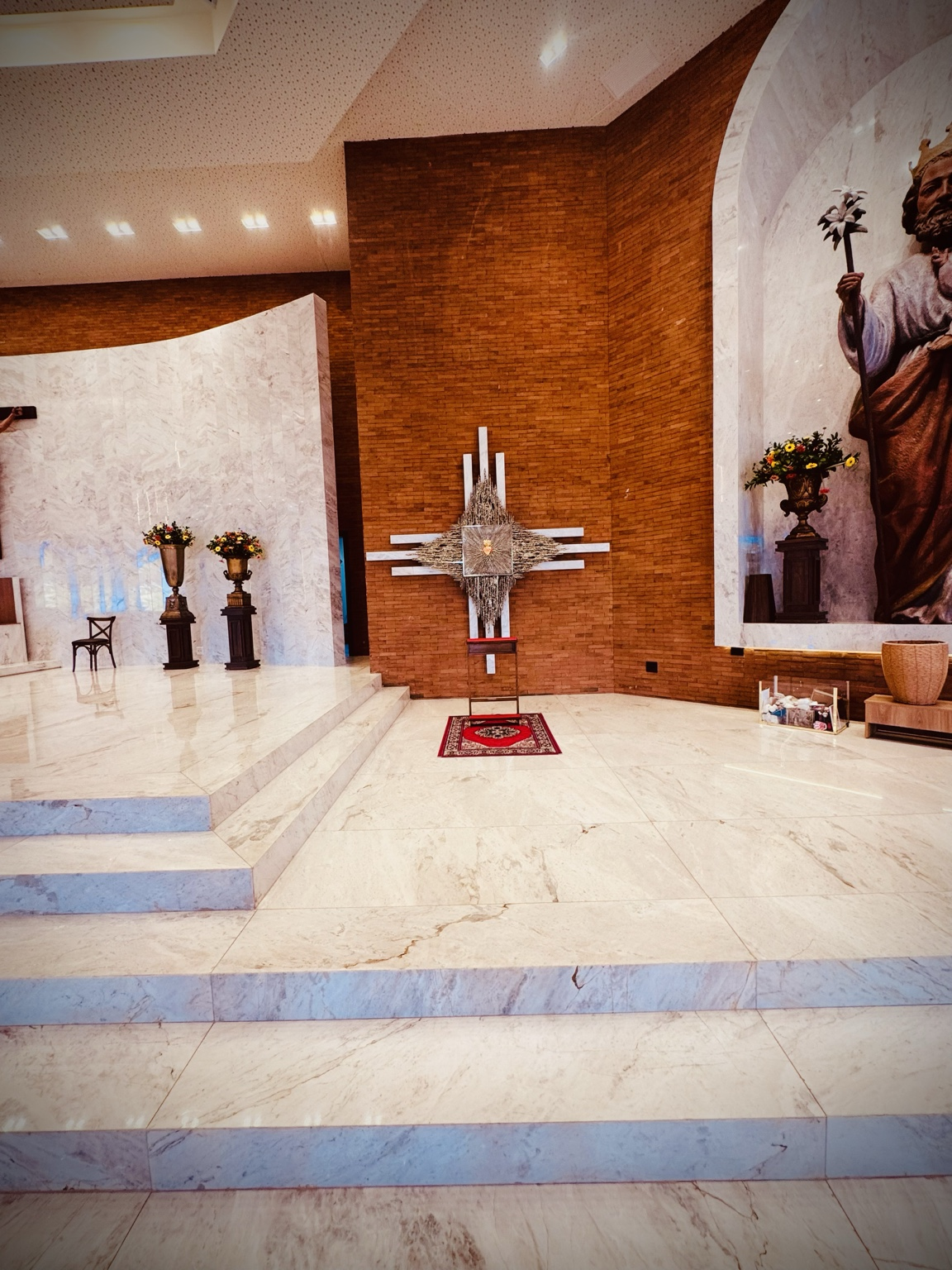 A fotografia apresenta um espaço de devoção localizado no interior do Santuário São José, em Alto Piquiri, Paraná. O registro destaca a integração entre arte sacra, arquitetura contemporânea e simbolismo religioso presentes na composição do ambiente. Ao centro da imagem encontra-se o painel artístico do Sagrado Coração de Jesus, instalado sobre uma cruz estilizada formada por elementos de mármore e estruturas metálicas ornamentais. A composição simboliza o amor, a misericórdia e a presença de Cristo na tradição cristã. Em frente ao painel observa-se um genuflexório destinado aos momentos de oração individual, reflexão e contemplação espiritual. O espaço foi projetado para favorecer a experiência de recolhimento dos visitantes e fiéis. À esquerda é possível visualizar parte do altar principal do santuário, revestido em mármore claro, enquanto à direita aparece parcialmente a escultura monumental de São José com o Menino Jesus, reforçando a identidade religiosa do templo e sua dedicação ao santo padroeiro. Os revestimentos em mármore, os painéis de tijolos aparentes e a iluminação cuidadosamente distribuída valorizam o conjunto arquitetônico e artístico, contribuindo para a formação de um ambiente de beleza, espiritualidade e acolhimento. Este espaço integra o patrimônio religioso, artístico e cultural do município de Alto Piquiri, constituindo importante elemento da memória e da identidade da comunidade local. Fonte: Registro fotográfico da equipe do Projeto EPT, 2026.