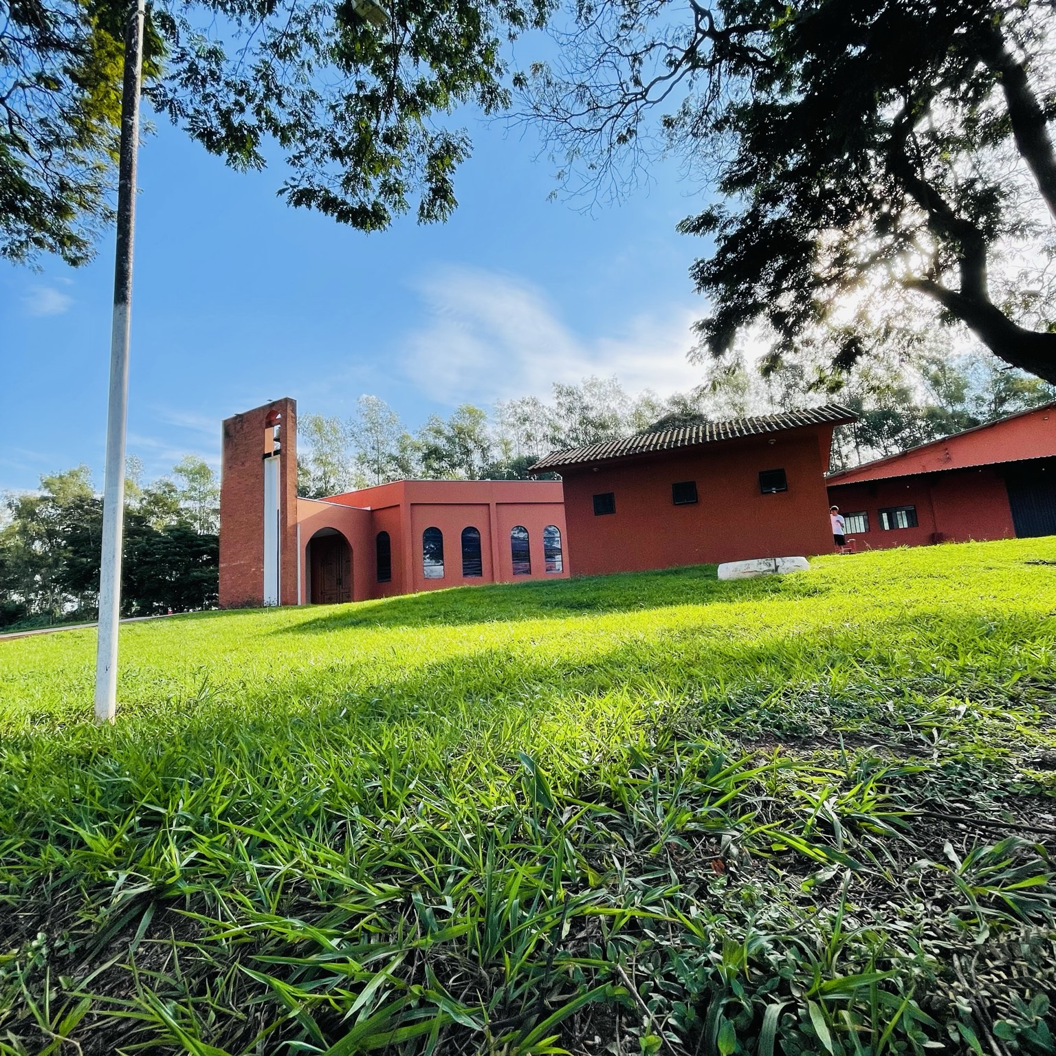 A fotografia apresenta uma vista externa da Capela Nossa Senhora Aparecida, localizada na comunidade de Mirante do Piquiri, área rural pertencente ao município de Alto Piquiri, no noroeste do Paraná. O registro destaca a integração harmoniosa entre a arquitetura religiosa e a paisagem natural que caracteriza a região. A capela encontra-se inserida em uma ampla área gramada, cercada por árvores de grande porte que proporcionam sombra, beleza paisagística e um ambiente propício à contemplação e à espiritualidade. A edificação possui linhas arquitetônicas simples e acolhedoras, características frequentemente encontradas nas capelas das comunidades do interior paranaense. Destaca-se a torre sineira construída em alvenaria, elemento tradicional da arquitetura religiosa cristã responsável por abrigar o sino utilizado para anunciar celebrações, encontros comunitários e momentos festivos da comunidade. A fachada apresenta portas e janelas em formato arqueado, recurso arquitetônico amplamente utilizado em edificações religiosas por remeter à tradição cristã e favorecer a entrada de luz natural nos espaços internos. Mais do que um local destinado às celebrações religiosas, a Capela Nossa Senhora Aparecida representa um importante patrimônio histórico, cultural e afetivo para os moradores do distrito de Mirante do Piquiri. Ao longo das décadas, o espaço tornou-se ponto de encontro comunitário, preservando tradições religiosas, manifestações culturais e memórias coletivas da população local. A devoção a Nossa Senhora Aparecida, padroeira do Brasil desde 1930, reforça a relevância simbólica do local, que permanece como referência de fé, identidade e pertencimento para diversas gerações da comunidade.