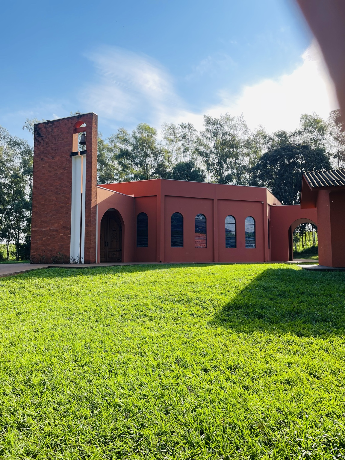A fotografia apresenta a fachada principal da Capela Nossa Senhora Aparecida, localizada na comunidade de Mirante do Piquiri, área rural do município de Alto Piquiri, no estado do Paraná. O registro destaca a arquitetura simples e acolhedora da capela, característica marcante dos templos religiosos construídos para atender as comunidades do interior brasileiro. Em primeiro plano observa-se uma ampla área gramada que valoriza a integração entre o espaço religioso e a paisagem natural que o cerca. À esquerda da composição encontra-se a torre sineira construída em tijolos aparentes, elemento tradicional da arquitetura cristã destinado a abrigar o sino utilizado para anunciar missas, celebrações religiosas e encontros comunitários. A presença da torre constitui um importante marco visual e simbólico da capela. O edifício apresenta linhas arquitetônicas sóbrias, com janelas em formato arqueado que favorecem a iluminação natural do ambiente interno. Os tons terrosos utilizados na construção harmonizam-se com a vegetação existente ao redor, criando uma paisagem que reforça o sentimento de tranquilidade e espiritualidade. Historicamente, as capelas rurais desempenharam papel fundamental na formação das comunidades do interior paranaense. Além de espaços destinados à oração e às celebrações religiosas, tornaram-se locais de encontro, convivência social e preservação das tradições culturais transmitidas entre gerações. Dedicada a Nossa Senhora Aparecida, padroeira do Brasil, a capela representa um importante símbolo de fé e identidade para os moradores de Mirante do Piquiri. Sua presença preserva a memória religiosa da comunidade e fortalece os laços culturais construídos ao longo do desenvolvimento da localidade. A Capela Nossa Senhora Aparecida integra o patrimônio religioso e cultural de Alto Piquiri, constituindo um espaço de devoção, acolhimento e valorização da história local. Fonte: Registro fotográfico da equipe do Projeto EPT, 2026.