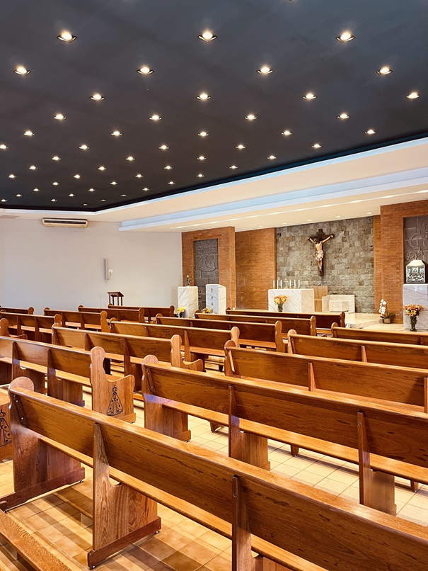 A fotografia apresenta o interior da Capela Nossa Senhora Aparecida, destacando a nave principal e o altar onde são realizadas as celebrações religiosas da comunidade de Mirante do Piquiri. O espaço é organizado por fileiras de bancos de madeira destinados aos fiéis, dispostos de forma a conduzir visualmente os visitantes em direção ao altar. Os bancos apresentam símbolos marianos gravados nas extremidades, reforçando a devoção a Nossa Senhora Aparecida, padroeira da capela e do Brasil. Ao fundo da imagem encontra-se o altar principal, construído em mármore claro e posicionado diante de um painel revestido em pedra natural. No centro destaca-se o crucifixo com a representação de Jesus Cristo, elemento central da fé cristã e referência para a liturgia católica. A iluminação distribuída uniformemente pelo teto cria um ambiente acolhedor e favorece a contemplação religiosa. A combinação entre madeira, pedra, mármore e elementos decorativos confere equilíbrio visual ao espaço, preservando a simplicidade característica das comunidades religiosas do interior paranaense. Historicamente, capelas como esta desempenham papel fundamental na vida das comunidades rurais, servindo não apenas como locais de celebração da fé, mas também como espaços de encontro, convivência social e preservação das tradições religiosas transmitidas entre gerações. A Capela Nossa Senhora Aparecida constitui importante patrimônio religioso e cultural de Mirante do Piquiri, preservando a memória da comunidade e fortalecendo os laços de identidade, espiritualidade e pertencimento dos moradores locais. Fonte: Registro fotográfico da equipe do Projeto EPT, 2026.Fonte: Registro fotográfico da equipe do Projeto EPT, 2026.