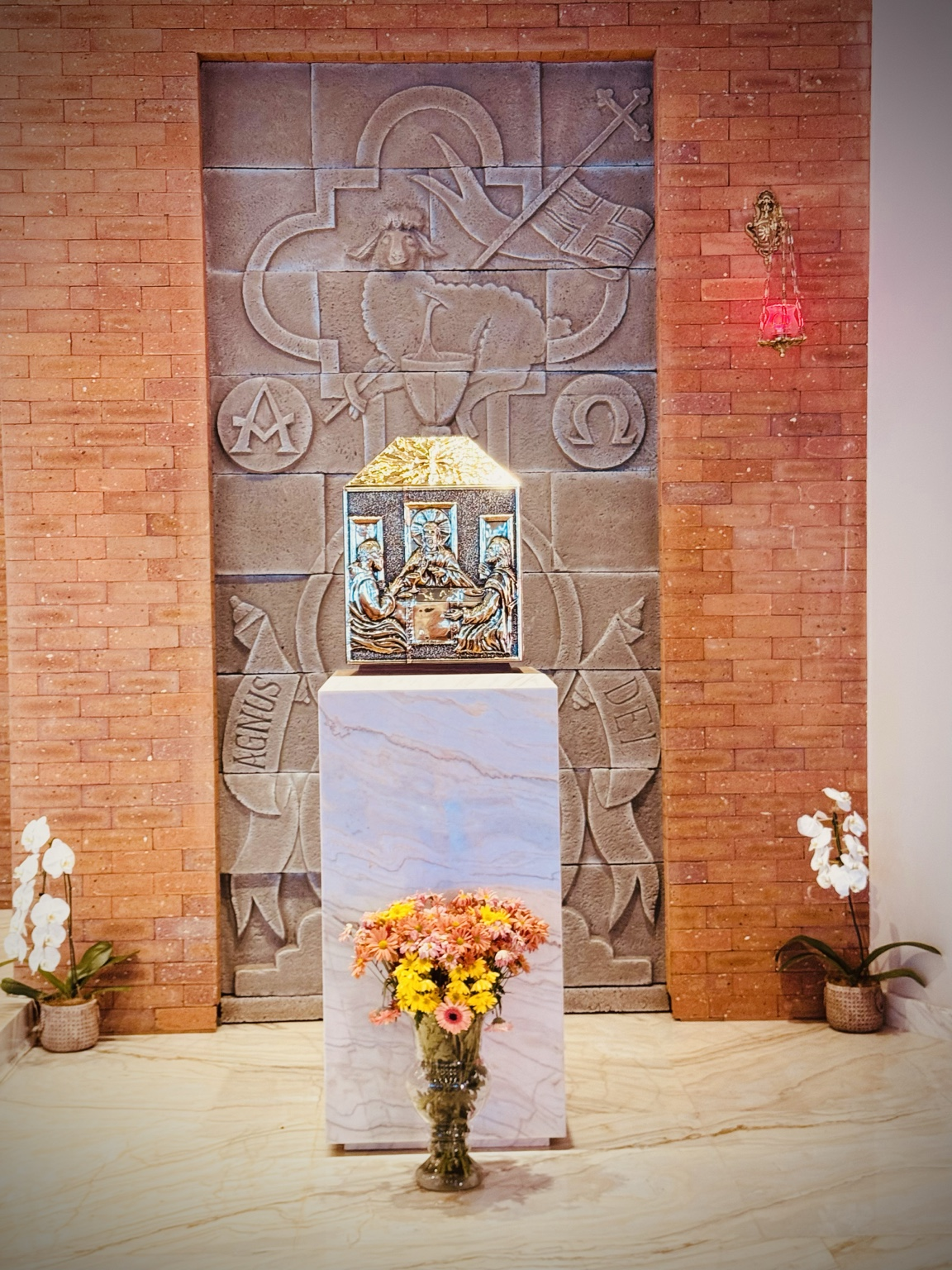A fotografia apresenta uma das laterais internas da Capela Nossa Senhora Aparecida, destacando a sequência de vitrais coloridos que acompanham a curvatura da nave. A composição valoriza a entrada da luz natural, que atravessa os painéis de vidro e projeta diferentes tonalidades sobre o ambiente religioso. Os vitrais possuem formas estilizadas e representam elementos da criação divina. Observam-se o céu azul, nuvens, águas, peixes, aves e áreas de vegetação, compondo uma paisagem inspirada na natureza. A obra dialoga com a tradição cristã que reconhece a criação como manifestação da presença de Deus no mundo. A predominância das cores azul, verde, branca e dourada cria um ambiente de contemplação e espiritualidade. A sequência dos vitrais acompanha a arquitetura da capela, conduzindo o olhar do visitante ao longo do espaço interno e reforçando a sensação de acolhimento e tranquilidade. Além de sua função decorativa, os vitrais possuem importante significado simbólico na tradição cristã. Historicamente, eles são utilizados para transmitir ensinamentos religiosos, valorizar a luz como representação do divino e enriquecer artisticamente os espaços de culto. Na Capela Nossa Senhora Aparecida, os vitrais integram o conjunto arquitetônico e artístico do templo, contribuindo para a preservação do patrimônio religioso e cultural da comunidade de Mirante do Piquiri. Fonte: Registro fotográfico da equipe do Projeto EPT, 2026.