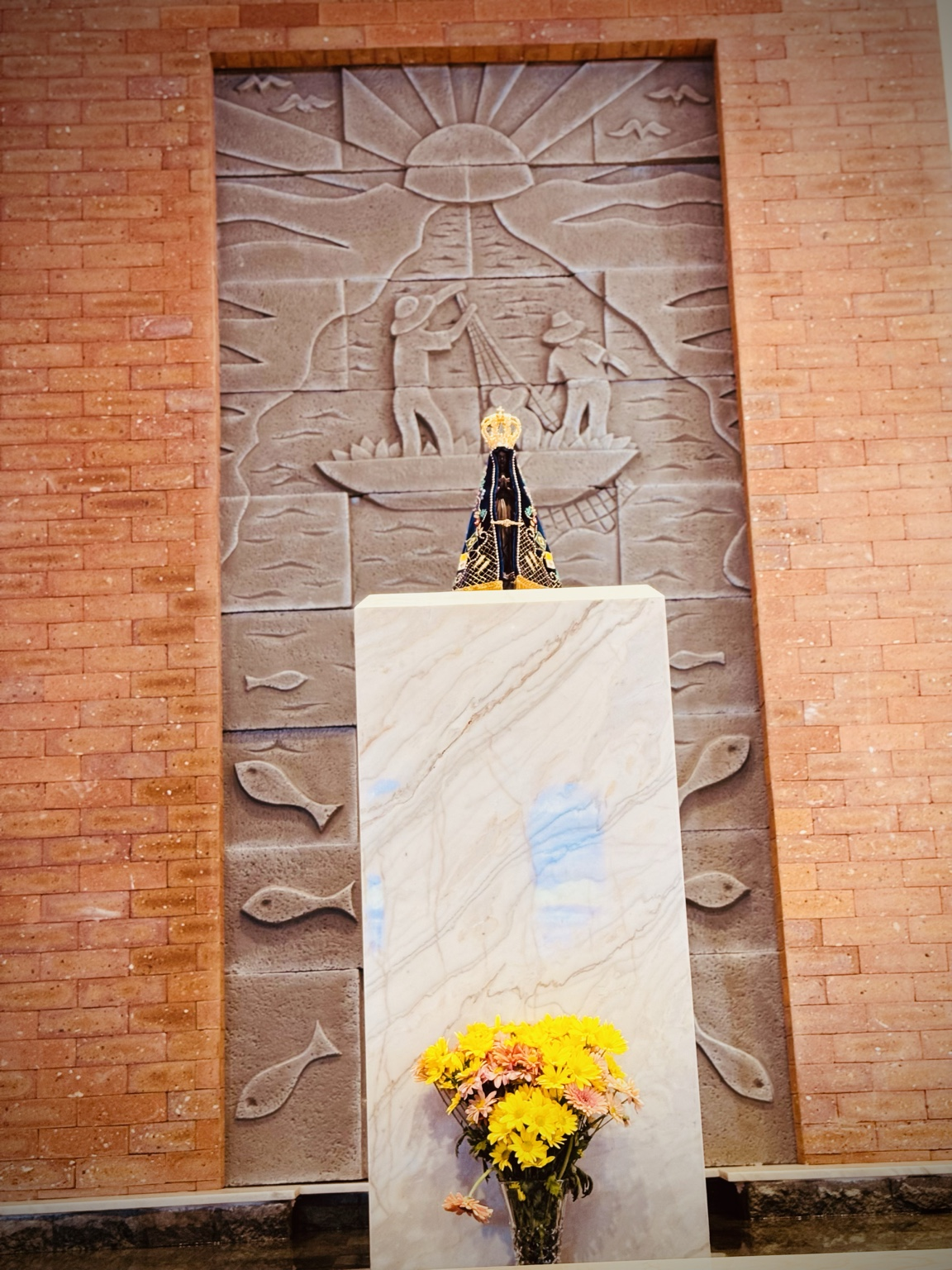 A fotografia registra uma área de grande importância litúrgica no interior da Capela Nossa Senhora Aparecida. Em destaque encontra-se o sacrário, estrutura destinada à guarda do Santíssimo Sacramento, posicionado sobre um pedestal revestido em mármore claro. Ao fundo observa-se um amplo painel artístico em relevo, composto por elementos simbólicos da tradição cristã. No centro destaca-se o Cordeiro de Deus, representação de Jesus Cristo sacrificado pela salvação da humanidade. Acima do cordeiro aparece a cruz processional e outros símbolos ligados à fé cristã. Nas laterais do painel encontram-se as letras gregas Alfa (Α) e Ômega (Ω), símbolos que representam Cristo como princípio e fim de todas as coisas, conforme descrito no livro do Apocalipse. O sacrário apresenta acabamento artístico em metal trabalhado, com cenas religiosas em relevo que reforçam seu valor devocional e patrimonial. A iluminação permanente da lâmpada vermelha, visível à direita da imagem, indica a presença do Santíssimo Sacramento, tradição mantida pela Igreja Católica em templos de todo o mundo.Na parte inferior observa-se um arranjo floral que contribui para o ambiente de acolhimento e oração. O conjunto arquitetônico harmoniza mármore, tijolos aparentes, esculturas e símbolos cristãos, compondo um espaço destinado à contemplação, ao silêncio e à espiritualidade. Além de seu valor religioso, este local integra o patrimônio cultural da comunidade de Mirante do Piquiri, preservando elementos artísticos que representam a história e a identidade da fé católica local. Fonte: Registro fotográfico da equipe do Projeto EPT, 2026