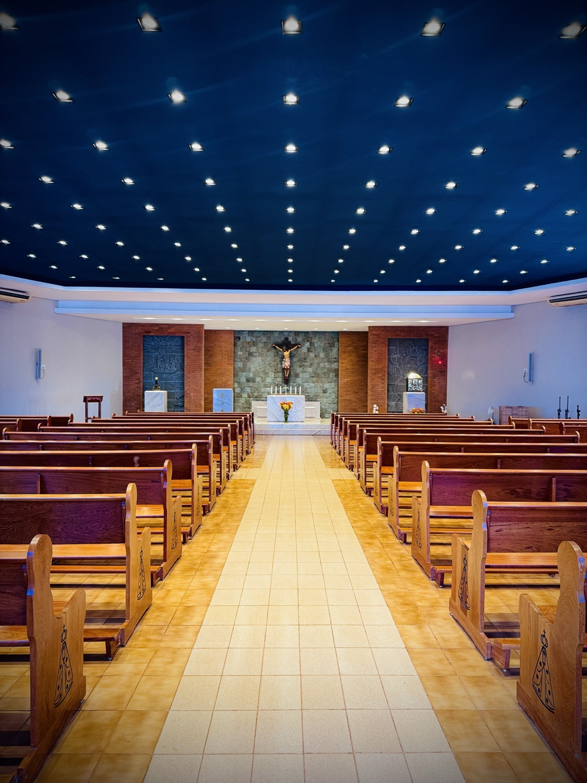 A fotografia apresenta uma vista central do interior da Capela Nossa Senhora Aparecida, permitindo observar a organização arquitetônica do espaço religioso a partir do corredor principal que conduz ao altar. O ambiente destaca-se pela simetria de suas linhas e pela disposição ordenada dos bancos de madeira, posicionados em ambos os lados da nave. Nas extremidades dos bancos observa-se a representação de Nossa Senhora Aparecida, reforçando a identidade religiosa da capela e a devoção da comunidade local à padroeira do Brasil. Ao fundo encontra-se o presbitério, espaço reservado às celebrações litúrgicas. Em posição central destaca-se o altar, construído em mármore claro, tendo ao fundo um grande crucifixo representando Jesus Cristo crucificado, principal símbolo da fé cristã. Nas laterais do altar podem ser observados espaços devocionais dedicados à religiosidade católica, integrando imagens, painéis artísticos e elementos simbólicos ligados à história da fé cristão teto chama atenção pelo acabamento escuro e pela distribuição uniforme da iluminação, que produz um efeito visual semelhante a um céu estrelado, contribuindo para a atmosfera de recolhimento, silêncio e contemplação. A arquitetura interna da capela combina simplicidade, funcionalidade e espiritualidade, valorizando a participação dos fiéis durante as celebrações religiosas e fortalecendo o papel da igreja como espaço de encontro comunitário, oração e preservação cultural. Além de seu significado religioso, a capela representa importante patrimônio histórico e cultural para a comunidade de Mirante do Piquiri, preservando tradições religiosas transmitidas ao longo das gerações. Fonte: Registro fotográfico da equipe do Projeto EPT, 2026.
