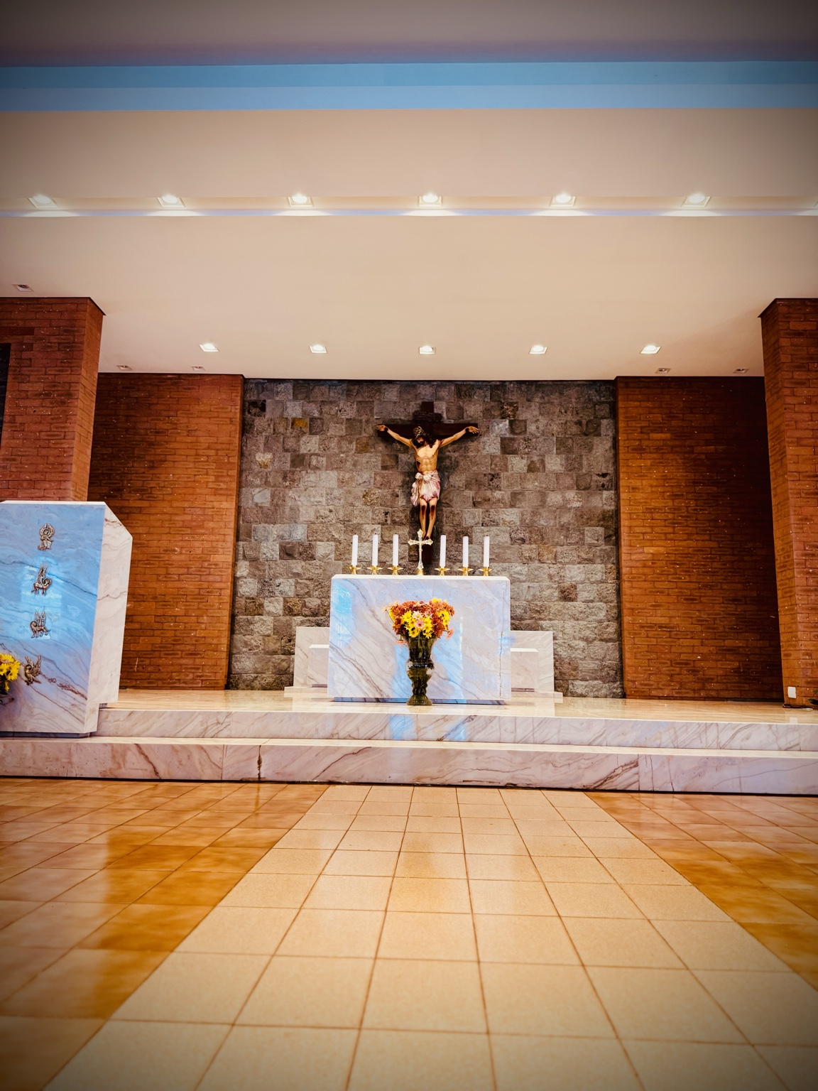 A fotografia apresenta o altar principal da Capela Nossa Senhora Aparecida, considerado o centro litúrgico e espiritual do templo. O registro foi realizado a partir da nave central, permitindo visualizar a composição arquitetônica que organiza o espaço destinado às celebrações religiosas. Em destaque encontra-se o altar em mármore claro, local onde são celebradas as missas e demais cerimônias da Igreja Católica. Sobre ele observa-se um arranjo floral que simboliza acolhimento, respeito e devoção. Ao fundo está o crucifixo com a imagem de Jesus Cristo crucificado, principal símbolo da fé cristã. A cruz recorda o sacrifício de Cristo e representa a esperança da ressurreição, fundamento da tradição cristã. A parede posterior é revestida por pedras naturais em diferentes tonalidades, criando um contraste harmonioso com o mármore do altar e valorizando visualmente o conjunto religioso. Nas laterais observam-se colunas revestidas em tijolos aparentes, elemento arquitetônico presente em diversos ambientes da capela. À esquerda da imagem encontra-se o ambão, local utilizado para a proclamação das leituras bíblicas e do Evangelho durante as celebrações. O conjunto foi planejado para favorecer a participação dos fiéis e a centralidade da liturgia. A simplicidade dos elementos arquitetônicos, associada à iluminação suave e aos materiais nobres utilizados na construção, transmite uma atmosfera de recolhimento, contemplação e espiritualidade. Além de sua função religiosa, o altar principal integra o patrimônio cultural e histórico da comunidade de Mirante do Piquiri, preservando tradições e manifestações de fé transmitidas ao longo das gerações. Fonte: Registro fotográfico da equipe do Projeto EPT, 2026.