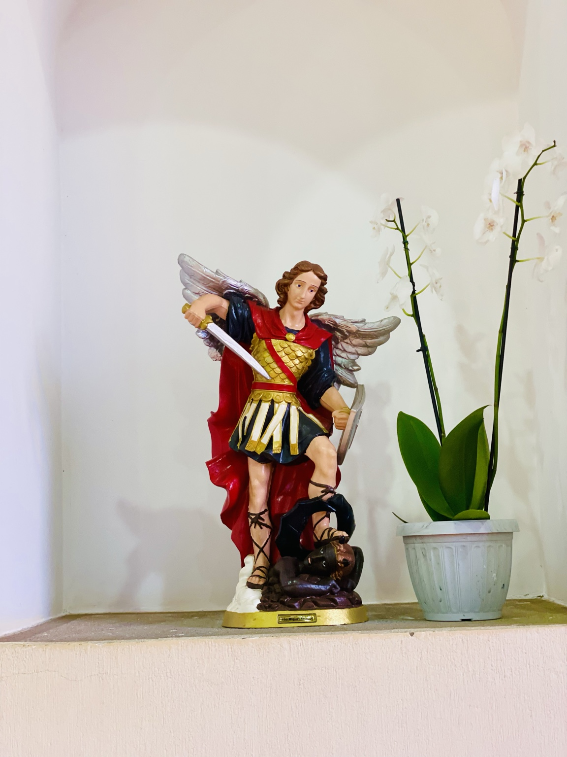 A fotografia apresenta uma imagem sacra de São Miguel Arcanjo posicionada em um nicho interno da Capela Santo Antônio. A escultura é colorida e representa o arcanjo em postura de combate e vitória sobre o mal. São Miguel aparece com grandes asas abertas, vestindo armadura dourada, túnica escura e manto vermelho que se destaca sobre o fundo claro da parede. Na mão direita, segura uma espada apontada para baixo, simbolizando a justiça divina e a proteção dos fiéis. Sob seus pés encontra-se a figura do mal derrotado, representada por uma criatura escura, reforçando a tradição cristã de São Miguel como defensor do bem. Ao lado da imagem há um vaso com orquídeas brancas, elemento decorativo que transmite serenidade e reverência ao ambiente religioso. A iluminação suave destaca os detalhes da escultura e cria uma atmosfera de contemplação e espiritualidade. Importância Histórico-Religiosa São Miguel Arcanjo é um dos santos mais venerados da tradição cristã. Seu nome significa “Quem como Deus?” e sua figura está associada à proteção, à coragem e à luta contra as forças do mal. Ao longo da história da Igreja Católica, São Miguel tornou-se símbolo da defesa da fé e da justiça divina. Sua presença em templos religiosos recorda aos visitantes valores como perseverança, fé, proteção espiritual e confiança em Deus. Na Capela Santo Antônio, a imagem integra o conjunto de arte sacra que contribui para a preservação da religiosidade local e da identidade cultural da comunidade de Paulistânia.