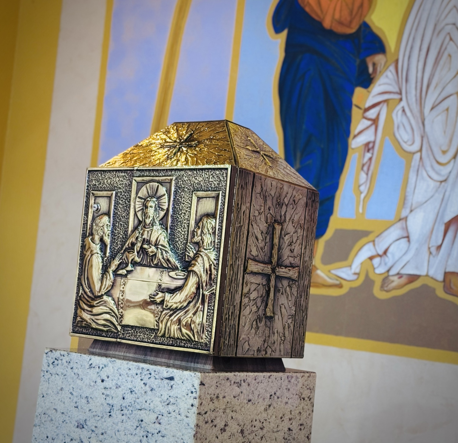 A fotografia apresenta o sacrário da Capela Santo Antônio em destaque. A peça possui formato arquitetônico semelhante a uma pequena capela ou templo, confeccionada em metal trabalhado com detalhes dourados e relevos religiosos. Na face frontal observa-se uma representação da Última Ceia, momento em que Jesus compartilha o pão e o vinho com seus discípulos. A cena está esculpida em alto relevo, valorizando a importância da Eucaristia para a fé cristã. Nas laterais do sacrário aparecem símbolos cristãos, incluindo a cruz, elemento central da espiritualidade católica. A peça encontra-se apoiada sobre um pedestal de pedra clara, recebendo destaque dentro do espaço litúrgico. Ao fundo, percebe-se parte de uma pintura religiosa em tons de azul, branco e dourado, compondo um ambiente harmonioso de oração e contemplação.
Importância Histórico-Religiosa
O sacrário é um dos elementos mais importantes de uma igreja católica. É nele que são guardadas as hóstias consagradas durante a celebração da missa, consideradas pelos fiéis a presença real de Cristo na Eucaristia.
A representação da Última Ceia recorda a instituição da Eucaristia realizada por Jesus Cristo na véspera de sua crucificação, evento fundamental para a tradição cristã.
Além de sua função litúrgica, o sacrário constitui importante expressão da arte sacra, reunindo simbolismo religioso, trabalho artesanal e patrimônio cultural preservado pela comunidade local.
Na Capela Santo Antônio, este elemento reforça a identidade religiosa da comunidade de Paulistânia e representa um espaço permanente de fé, oração e devoção.