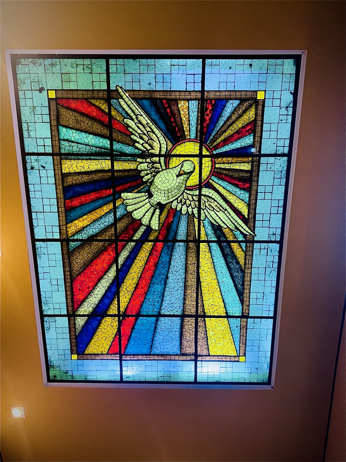 A fotografia registra um vitral artístico instalado no teto da Capela Santo Antônio. A composição apresenta a figura de uma pomba branca com as asas abertas, símbolo tradicional do Espírito Santo na fé cristã. Ao redor da pomba, raios coloridos em tons de azul, vermelho, amarelo e dourado irradiam-se a partir de um círculo luminoso central, criando um efeito visual que transmite movimento, luz e espiritualidade. O conjunto é formado por pequenas peças coloridas organizadas em mosaico, valorizando a técnica tradicional dos vitrais religiosos. A iluminação natural atravessa o vitral e projeta suas cores pelo interior da capela, contribuindo para a atmosfera de recolhimento, oração e contemplação.
Importância Histórico-Religiosa
Na tradição cristã, a pomba representa o Espírito Santo, terceira pessoa da Santíssima Trindade. Esse símbolo aparece em diversos momentos da história bíblica, especialmente no Batismo de Jesus, quando o Espírito Santo desceu sobre Ele em forma de pomba.
Os vitrais são elementos muito presentes na arquitetura religiosa desde a Idade Média. Além de sua função decorativa, servem para transmitir ensinamentos da fé por meio de imagens e símbolos facilmente compreendidos pelos fiéis.
Na Capela Santo Antônio, este vitral une arte, espiritualidade e patrimônio cultural, contribuindo para a identidade religiosa da comunidade de Paulistânia.