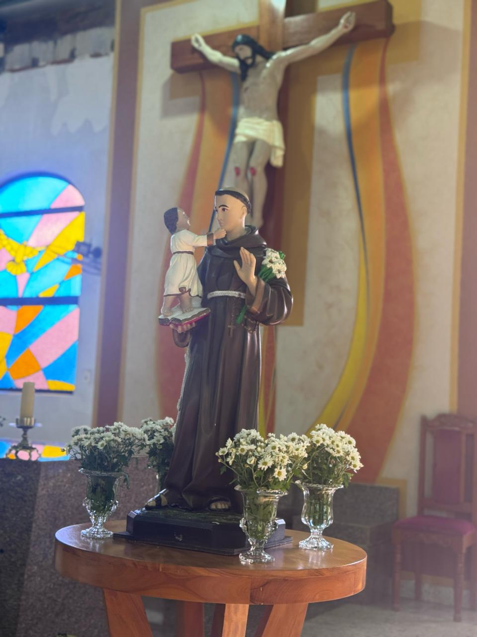 A fotografia apresenta a imagem de Santo Antônio de Pádua, posicionada sobre uma mesa de madeira no interior da Capela Santo Antônio. O santo é representado vestindo o tradicional hábito franciscano marrom, símbolo de humildade e serviço. Em seus braços encontra-se o Menino Jesus, uma das representações mais conhecidas da iconografia antoniana. Ao redor da imagem, vasos com flores brancas ornamentam o espaço, reforçando a atmosfera de devoção e acolhimento. Ao fundo, observa-se um crucifixo e vitrais coloridos que iluminam suavemente o ambiente, criando um cenário de fé, oração e contemplação. Contexto Histórico e Religioso Santo Antônio de Pádua nasceu em Lisboa, Portugal, no século XII, e tornou-se um dos santos mais populares da Igreja Católica. Conhecido por sua profunda dedicação aos ensinamentos cristãos e por sua atuação junto aos mais necessitados, é frequentemente invocado como intercessor das famílias e protetor daqueles que buscam auxílio espiritual. Sua devoção chegou ao Brasil com os colonizadores portugueses e permanece viva em inúmeras comunidades religiosas. No Distrito de Paulistânia, a Capela Santo Antônio representa um importante espaço de encontro, fé e preservação das tradições religiosas locais. A imagem retratada simboliza não apenas a devoção ao santo padroeiro, mas também a história de uma comunidade que mantém viva sua identidade cultural e espiritual através das geração.